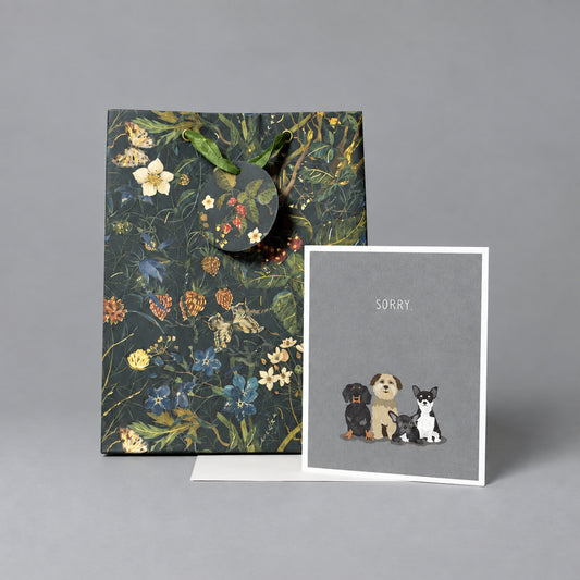 A floral gift bag with dark handles stands next to a greeting card that says 'SORRY' and shows small illustrations of three dogs, set against a textured light gray wall and concrete floor.