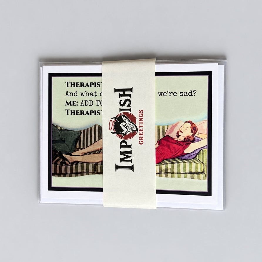 A stack of greeting cards wrapped in a cream paper band labeled 'Impish Greetings,' placed on a textured gray surface. The card front shows vintage-style illustrations and text including 'Therapy And what ( MG 1897) Therapy we're sad?'.