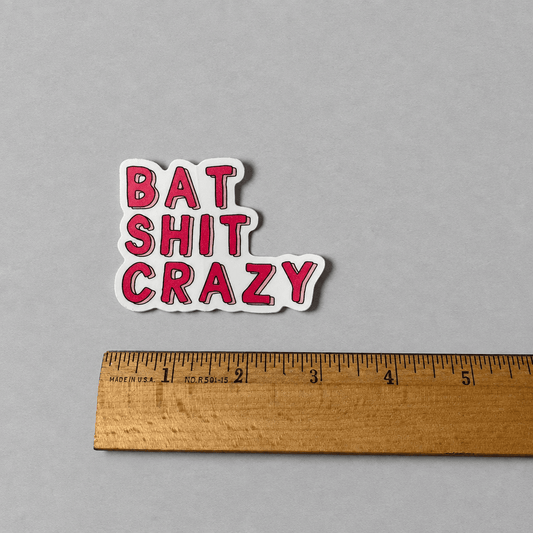 A die-cut vinyl sticker with the text 'BAT SHIT CRAZY' in bold pink letters outlined in black, with a white border, shown next to a wooden ruler on a neutral gray background.
