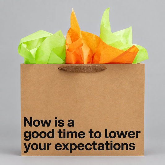 A brown kraft paper gift bag with black printed text reading 'Now is a good time to lower your expectations,' filled with bright orange and lime green tissue paper, on a light gray background.
