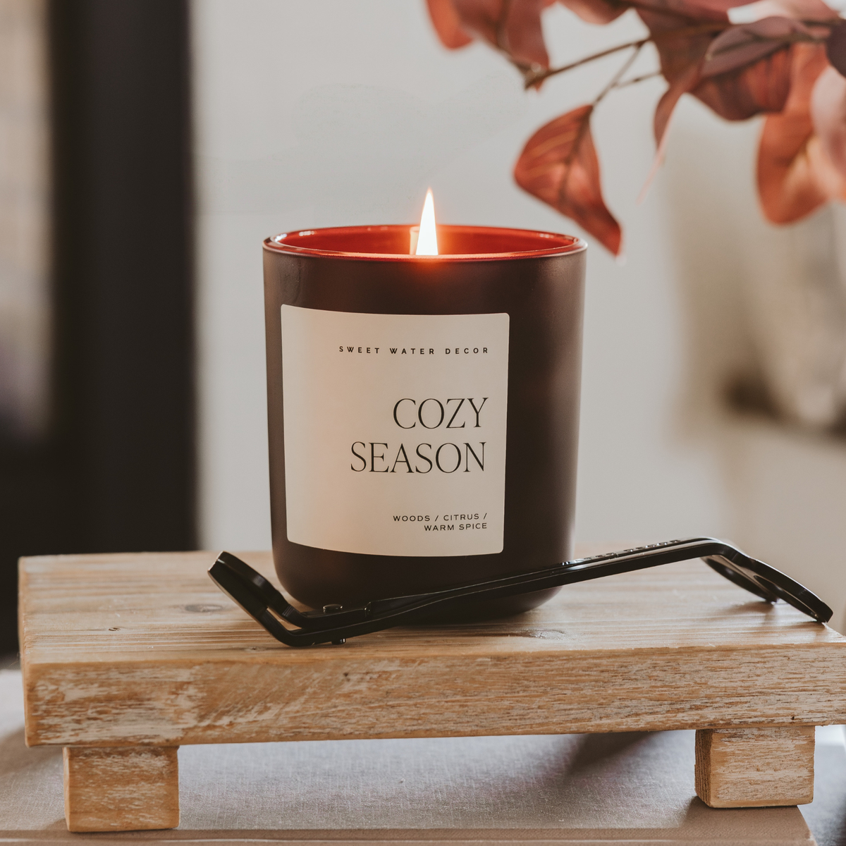 Impish Greetings 15 oz Cozy Season candle in a matte black jar with a lit wick, resting on a rustic wooden riser.