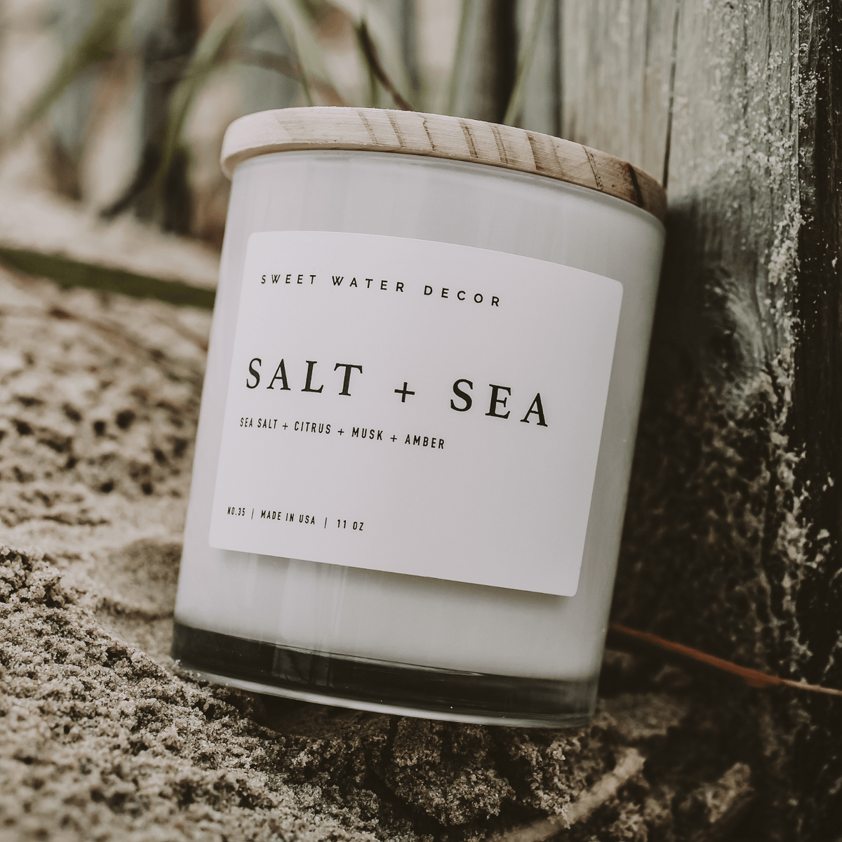 Impish Greetings Salt and Sea soy candle in a glass jar with a wooden lid, placed on sandy beach ground.