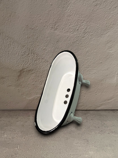 A vintage-style miniature white enamel bathtub with a black rim and light green legs, photographed against a textured gray concrete wall and floor.