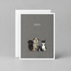 A greeting card with a dark gray background featuring three small illustrated dogs and the word 'SORRY.' printed above them, shown with a white envelope behind it on a textured gray surface.