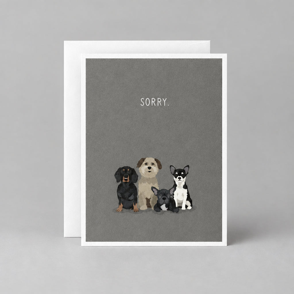 A greeting card with a dark gray background featuring three small illustrated dogs and the word 'SORRY.' printed above them, shown with a white envelope behind it on a textured gray surface.