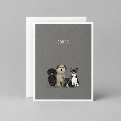 A greeting card with a dark gray background featuring three small illustrated dogs and the word 'SORRY.' printed above them, shown with a white envelope behind it on a textured gray surface.