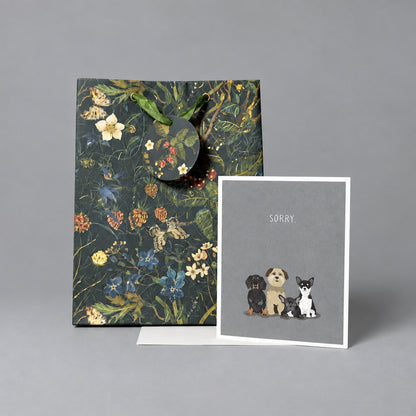 A floral gift bag with dark handles stands next to a greeting card that says 'SORRY' and shows small illustrations of three dogs, set against a textured light gray wall and concrete floor.