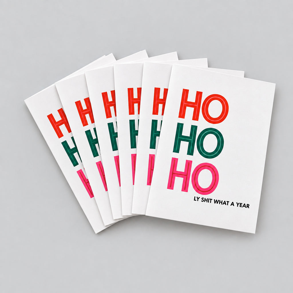 A set of white greeting cards stacked and fanned out on a textured gray concrete surface. The cards feature the phrase 'HO HO HO' in alternating red and green lettering, with the tagline 'LY SHIT WHAT A YEAR' printed in black on the bottom right of the front card.