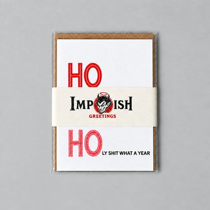 A stack of greeting cards from Impish Greetings featuring a bold red and black holiday design, wrapped in a beige paper band, placed on a textured gray concrete surface.