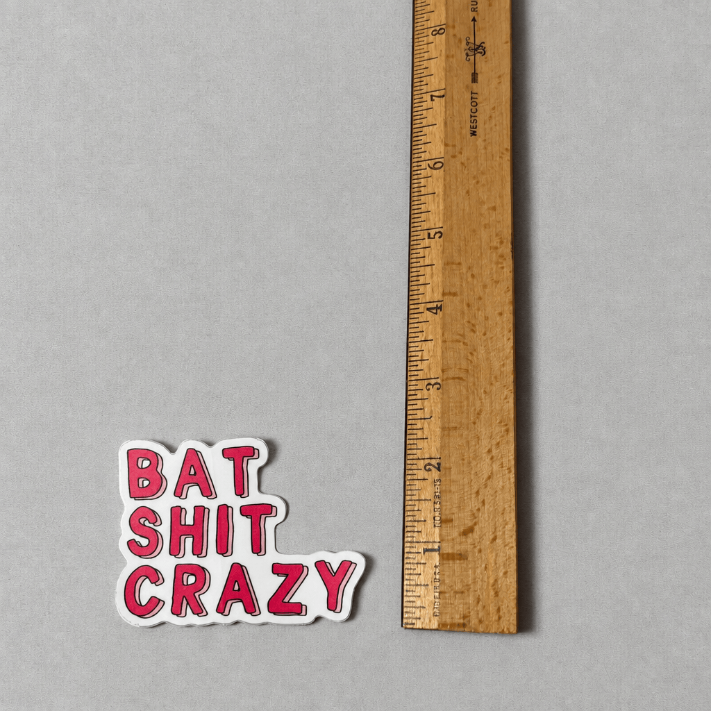 A die-cut vinyl sticker with the text 'BAT SHIT CRAZY' in red bubble letters outlined in black on a white background, shown next to a wooden ruler on a gray textured surface.