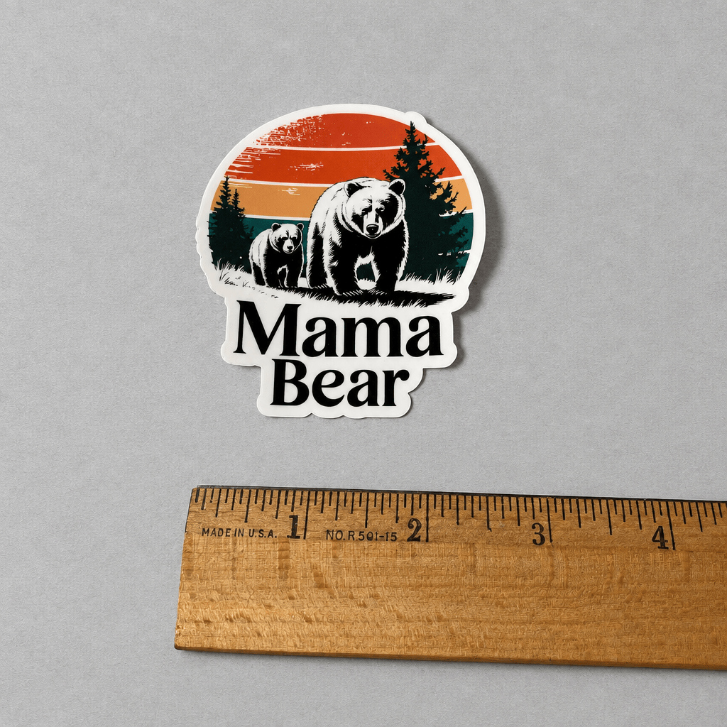 A die-cut vinyl sticker featuring a retro-style illustration of a mama bear and cub with pine trees and a sunset, placed above a wooden ruler on a neutral gray background.