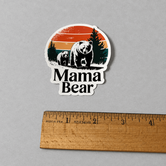 A die-cut vinyl sticker featuring a retro-style illustration of a mama bear and cub with pine trees and a sunset, placed above a wooden ruler on a neutral gray background.