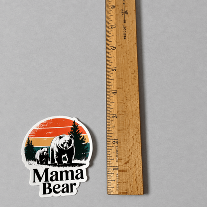 A die-cut sticker featuring a black and white mama bear and baby bear illustration with a retro orange, yellow, and blue sunset background and the text 'Mama Bear'. The sticker is displayed next to a wooden ruler on a neutral gray background to show its size.