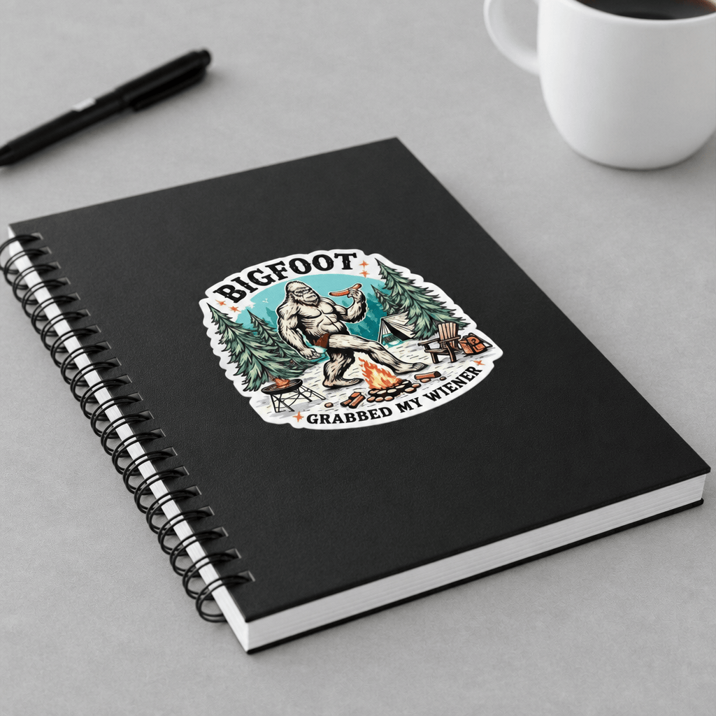 A black spiral notebook with a Bigfoot camping graphic sticker, shown on a gray tabletop with a black pen and a white mug in the background.