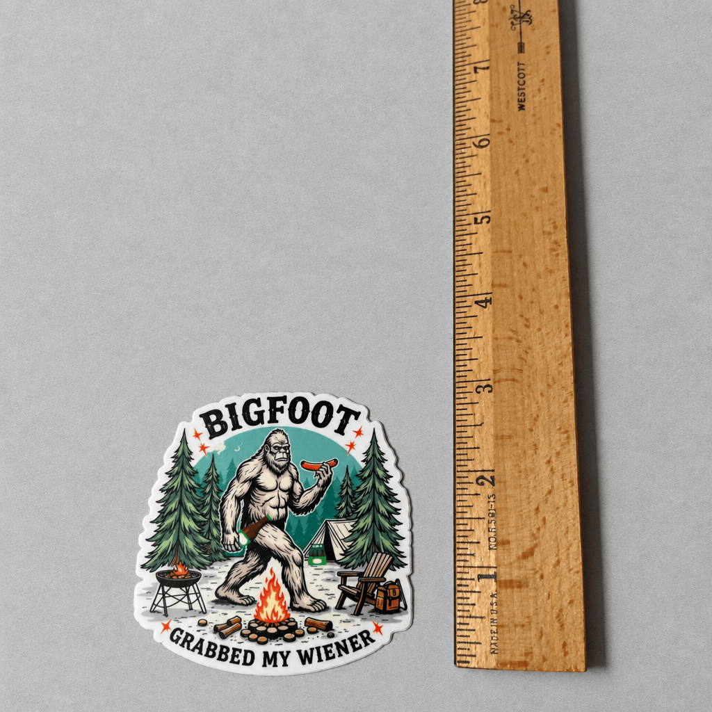 A die-cut sticker featuring a cartoon Bigfoot sitting by a campfire and holding a hot dog, with the text 'BIGFOOT' above and 'GRABBED MY WIENER' below. The sticker is displayed on a neutral gray background next to a wooden ruler for scale.