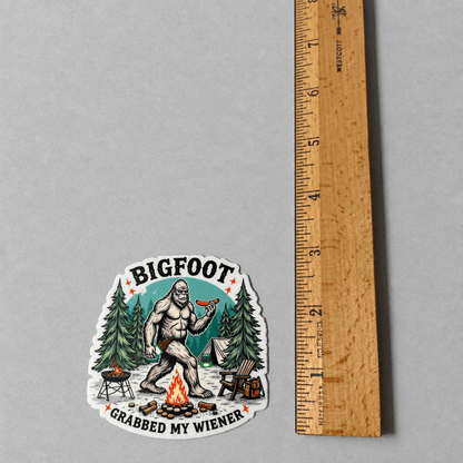 A die-cut sticker featuring a cartoon Bigfoot sitting by a campfire and holding a hot dog, with the text 'BIGFOOT' above and 'GRABBED MY WIENER' below. The sticker is displayed on a neutral gray background next to a wooden ruler for scale.