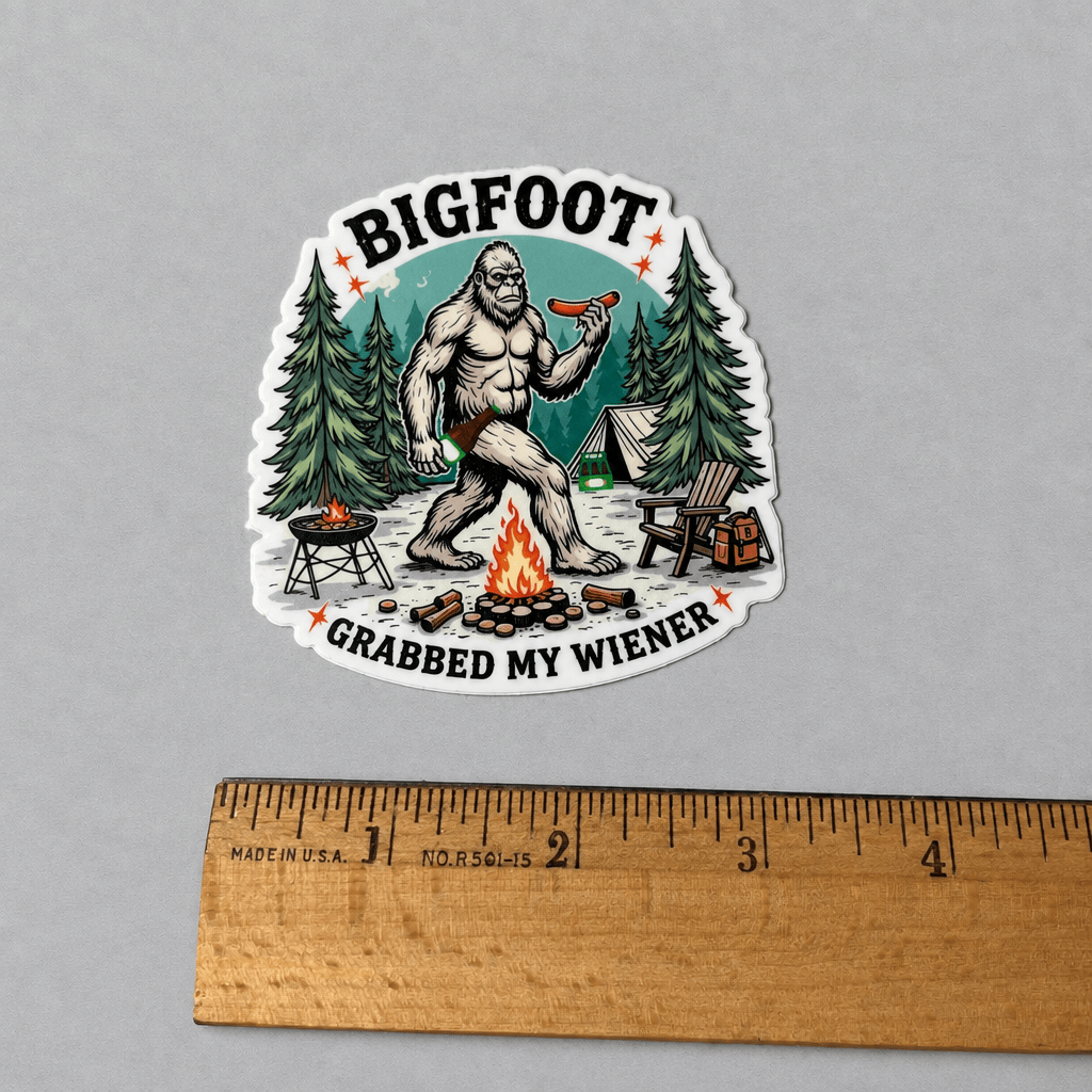 A die-cut sticker featuring a cartoon Bigfoot sitting by a campfire and holding a hot dog, with the text 'BIGFOOT' at the top and 'GRABBED MY WIENER' at the bottom. The sticker is displayed on a neutral gray background with a wooden ruler at the bottom for scale.