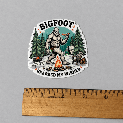 A die-cut sticker featuring a cartoon Bigfoot sitting by a campfire and holding a hot dog, with the text 'BIGFOOT' at the top and 'GRABBED MY WIENER' at the bottom. The sticker is displayed on a neutral gray background with a wooden ruler at the bottom for scale.
