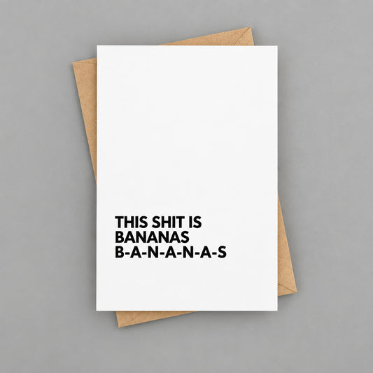 A white greeting card with bold black text reading 'THIS SHIT IS BANANAS B-A-N-A-N-A-S' stacked vertically, placed on top of a kraft paper envelope against a white wood plank background.