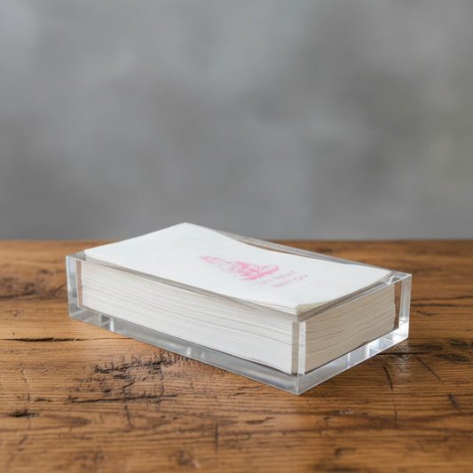 Impish Greetings paper guest towels with pink foil Buddha design in a clear acrylic holder on a wooden table.