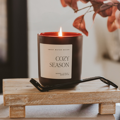 Impish Greetings 15 oz Cozy Season candle in a matte black jar with a lit wick, resting on a rustic wooden riser.