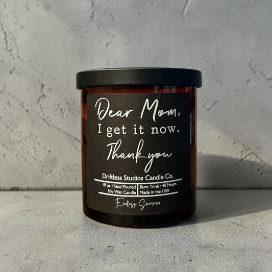 Impish Greetings Dear Mom I Get It Now soy wax candle in an amber glass jar with a black tin lid and white label.