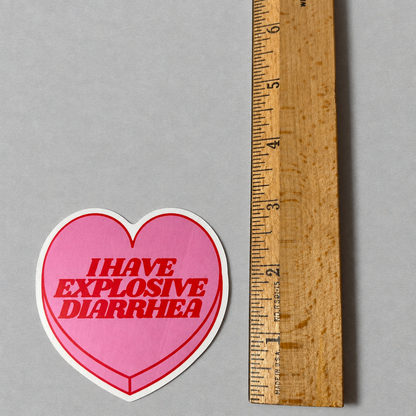 Impish Greetings heart-shaped pink vinyl sticker with red text reading I HAVE EXPLOSIVE DIARRHEA next to a ruler.