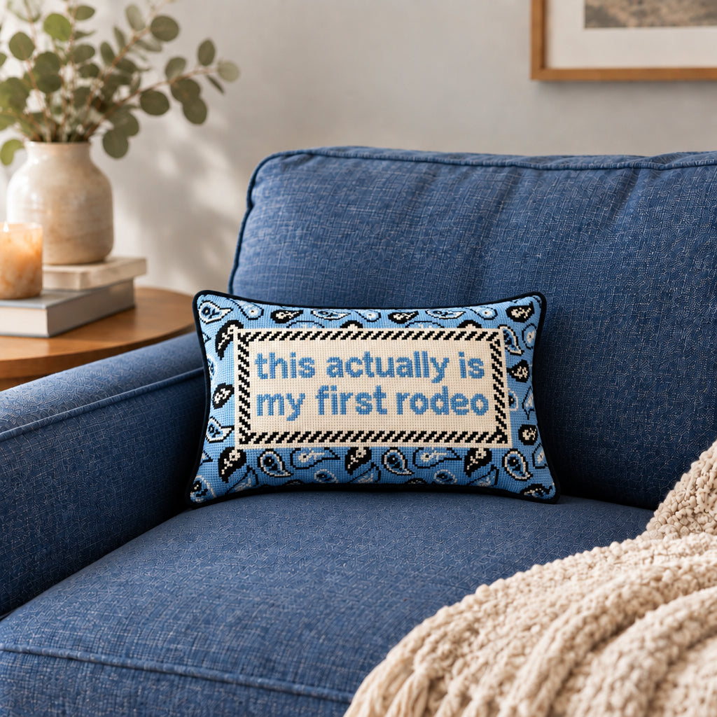 Impish Greetings First Rodeo needlepoint pillow with blue paisley pattern and witty text on a blue sofa