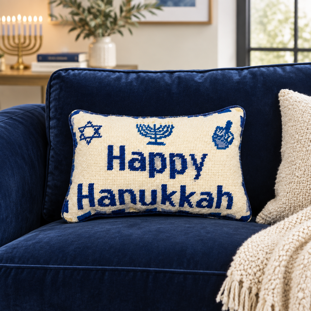 Impish Greetings Happy Hanukkah needlepoint pillow with blue menorah and dreidel motifs on a navy velvet sofa.