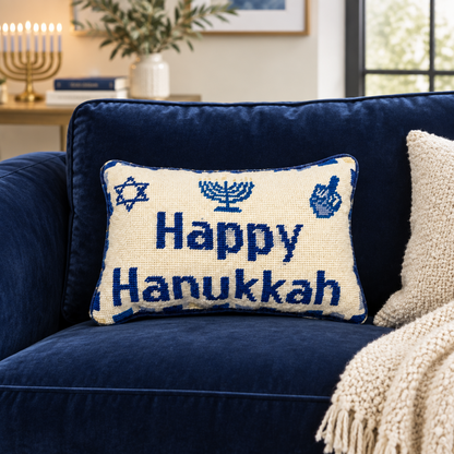 Impish Greetings Happy Hanukkah needlepoint pillow with blue menorah and dreidel motifs on a navy velvet sofa.