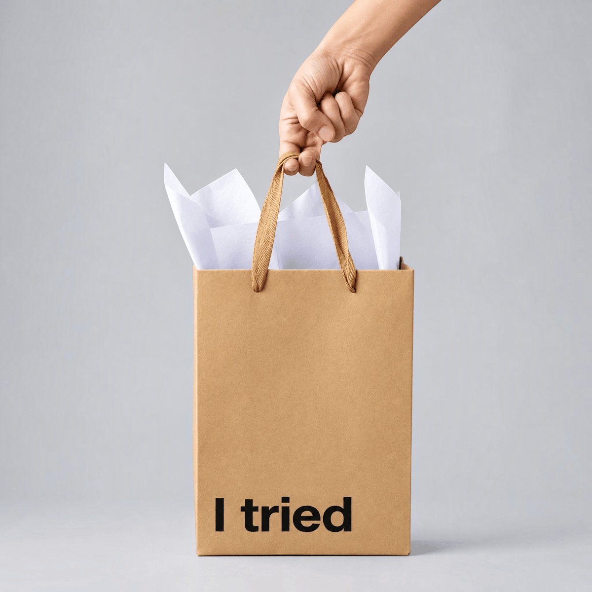 Impish Greetings I Tried kraft paper gift bag with ribbon handles held by a hand against a grey background