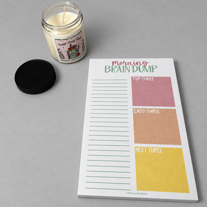 Impish Greetings Morning Brain Dump notepad with colorful task boxes and a scented candle on a grey surface.