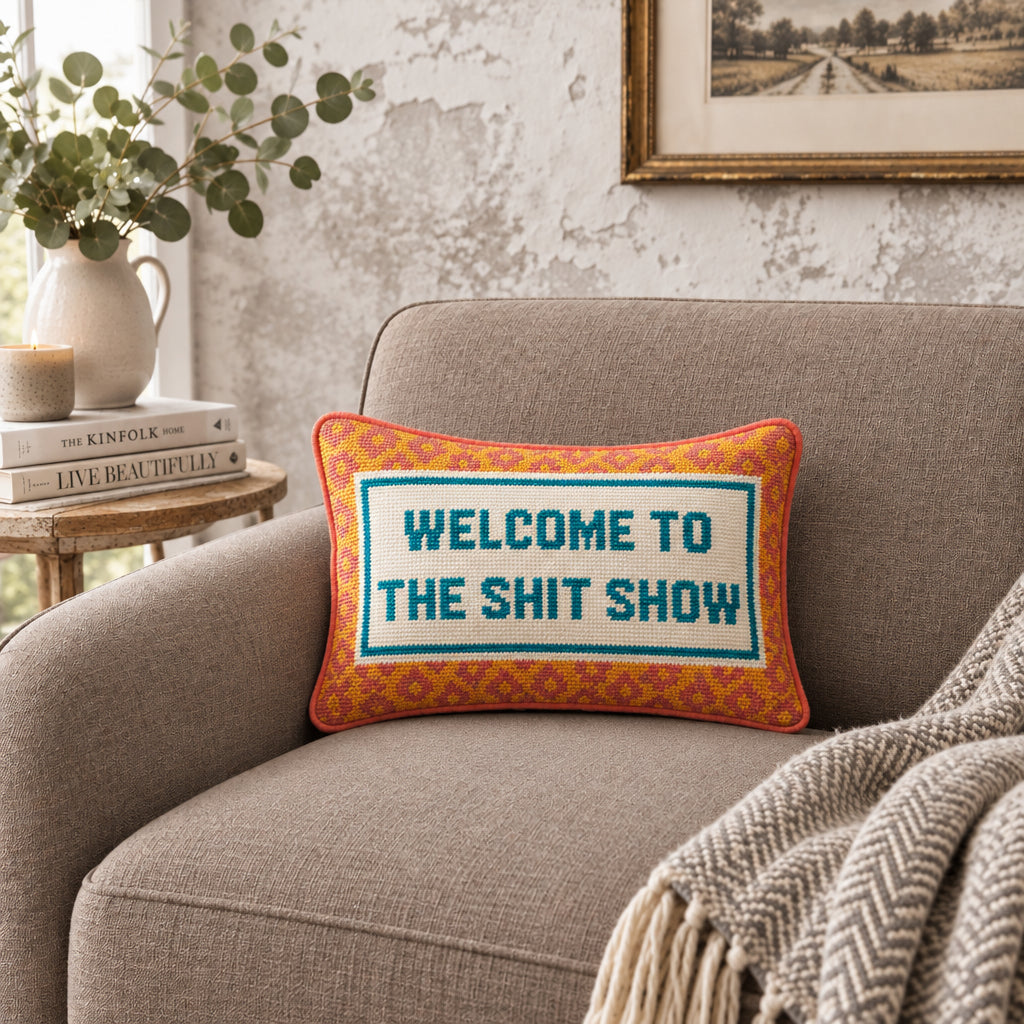 Impish Greetings needlepoint throw pillow with Welcome To The Shit Show text on a grey armchair