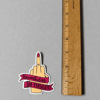 Impish Greetings Nevertheless She Persisted vinyl sticker featuring a hand giving the middle finger on a grey surface