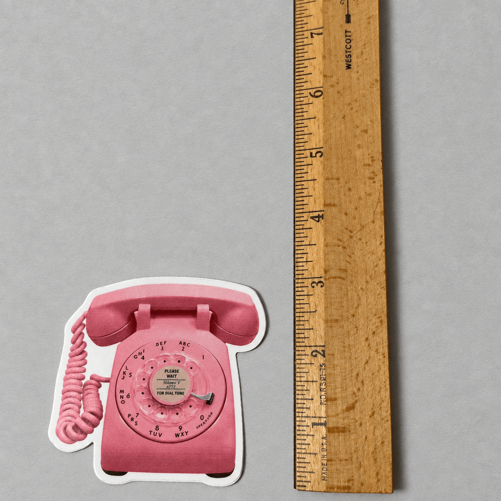 Impish Greetings retro pink rotary phone vinyl sticker next to a wooden ruler for scale on a grey background.