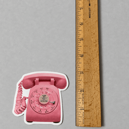 Impish Greetings retro pink rotary phone vinyl sticker next to a wooden ruler for scale on a grey background.