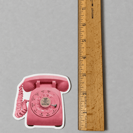 Impish Greetings retro pink rotary phone vinyl sticker next to a wooden ruler for scale on a grey background.