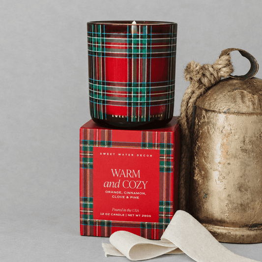 Impish Greetings Warm and Cozy soy candle in a red plaid glass jar with matching gift box and rustic metal bell.