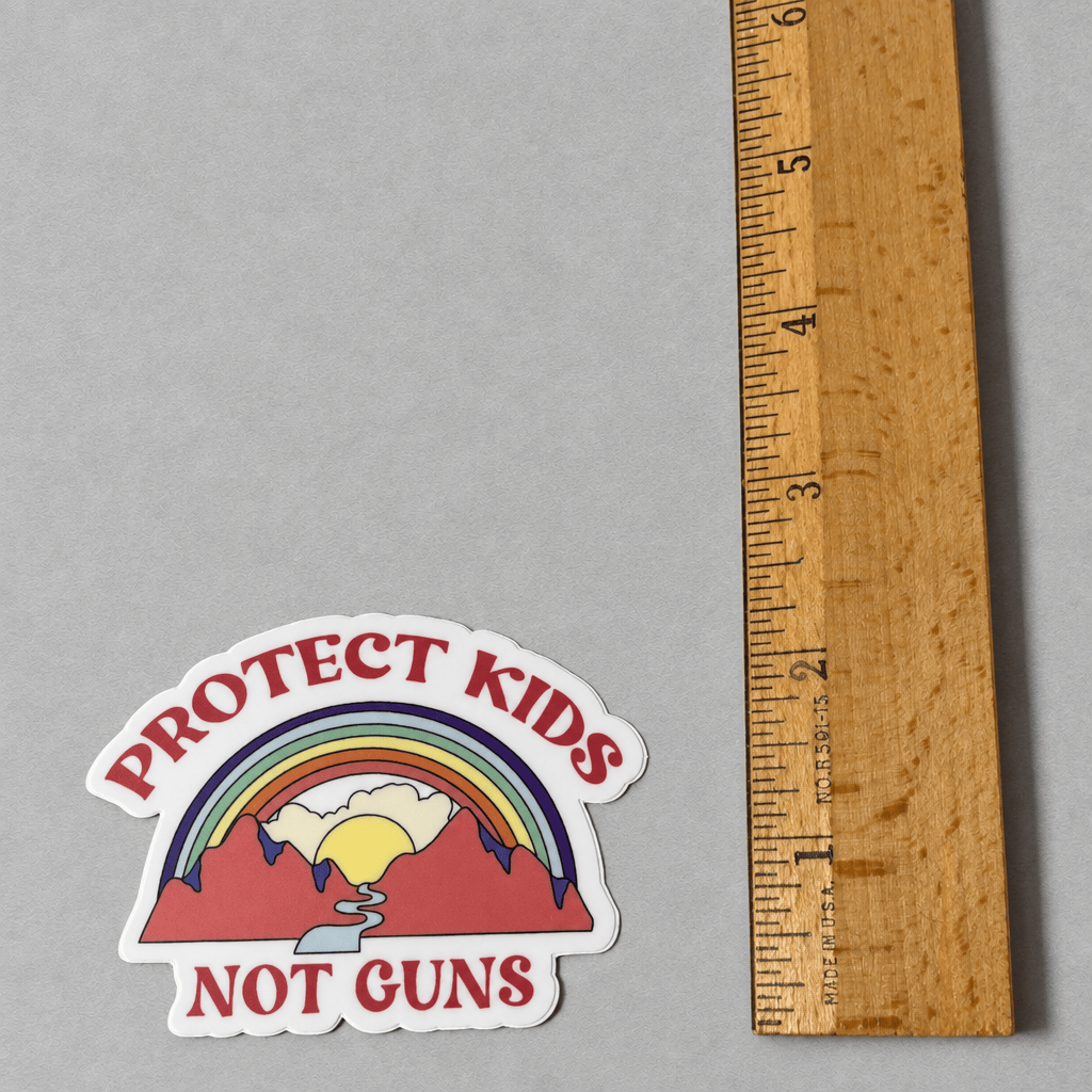 Impish Greetings Protect Kids Not Guns vinyl sticker with a rainbow and mountain design next to a wooden ruler.