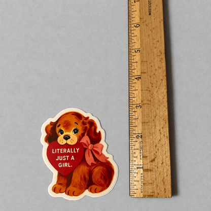 Impish Greetings Literally Just a Girl puppy sticker with a red heart and bow, shown next to a wooden ruler.