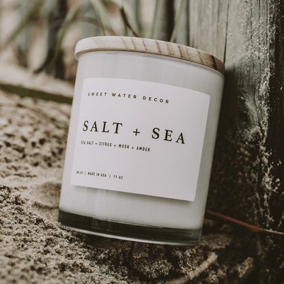 Impish Greetings Salt and Sea soy candle in a glass jar with a wooden lid, placed on sandy beach ground.