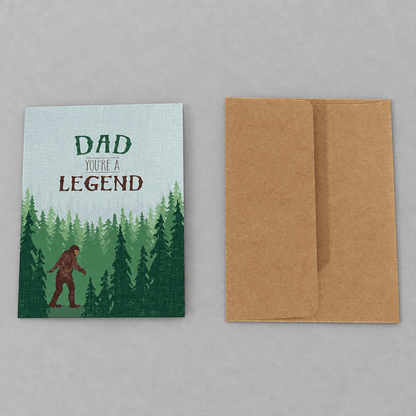 Impish Greetings Sasquatch Legend Dad card with forest illustration and brown kraft envelope on a grey background.