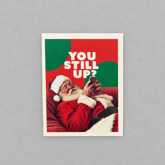 Impish Greetings Still Up? Christmas card featuring Santa Claus lying on a red sofa looking at his smartphone.
