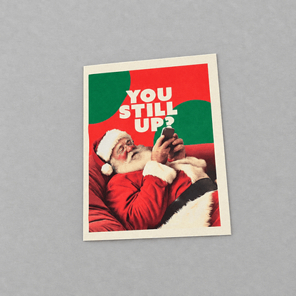 Impish Greetings Still Up? Christmas card featuring Santa Claus looking at his smartphone on a red background.